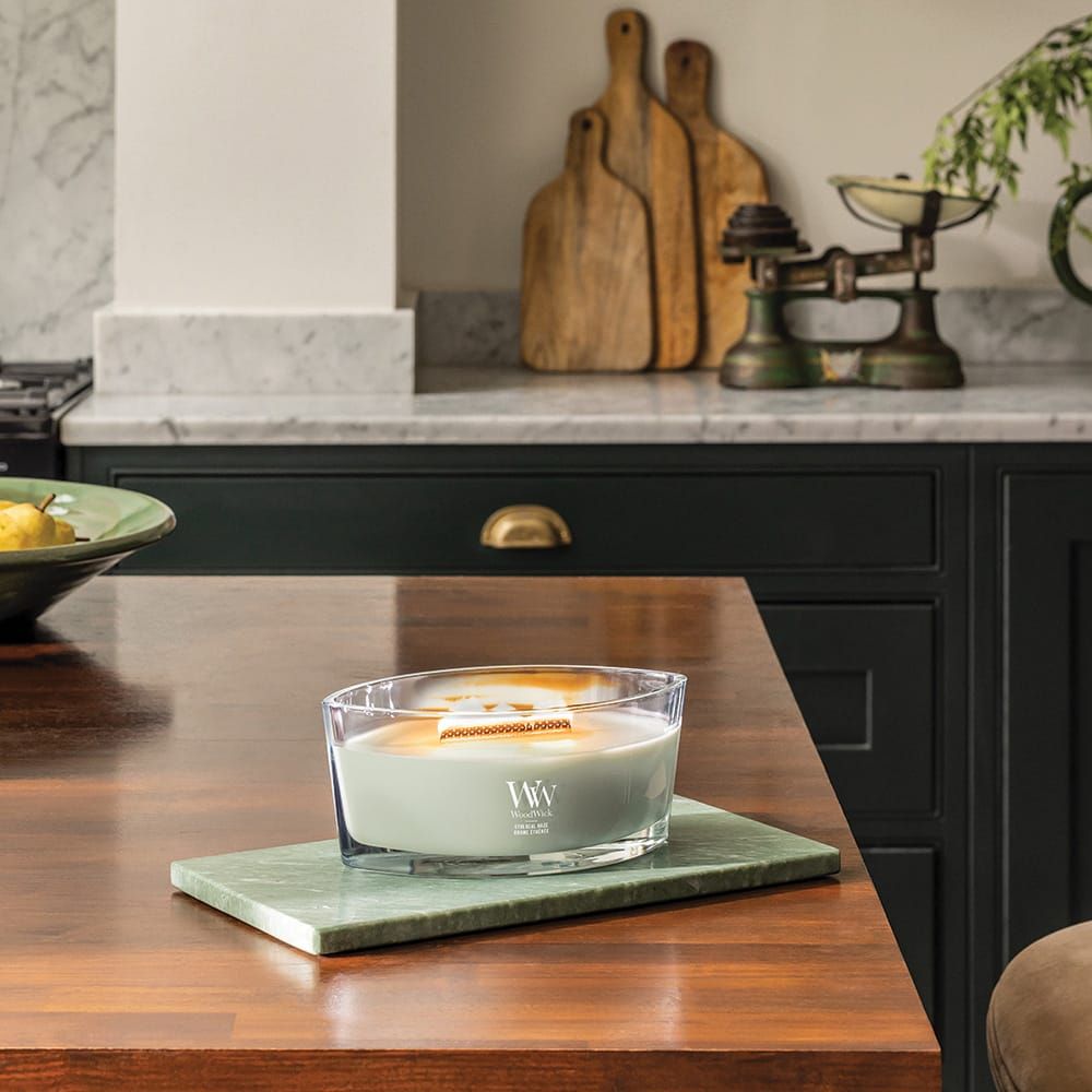 Candela con stoppino in legno su piastra verde. Contenitore in vetro, cera verde. Logo 'WW'. Su tavolo di legno.