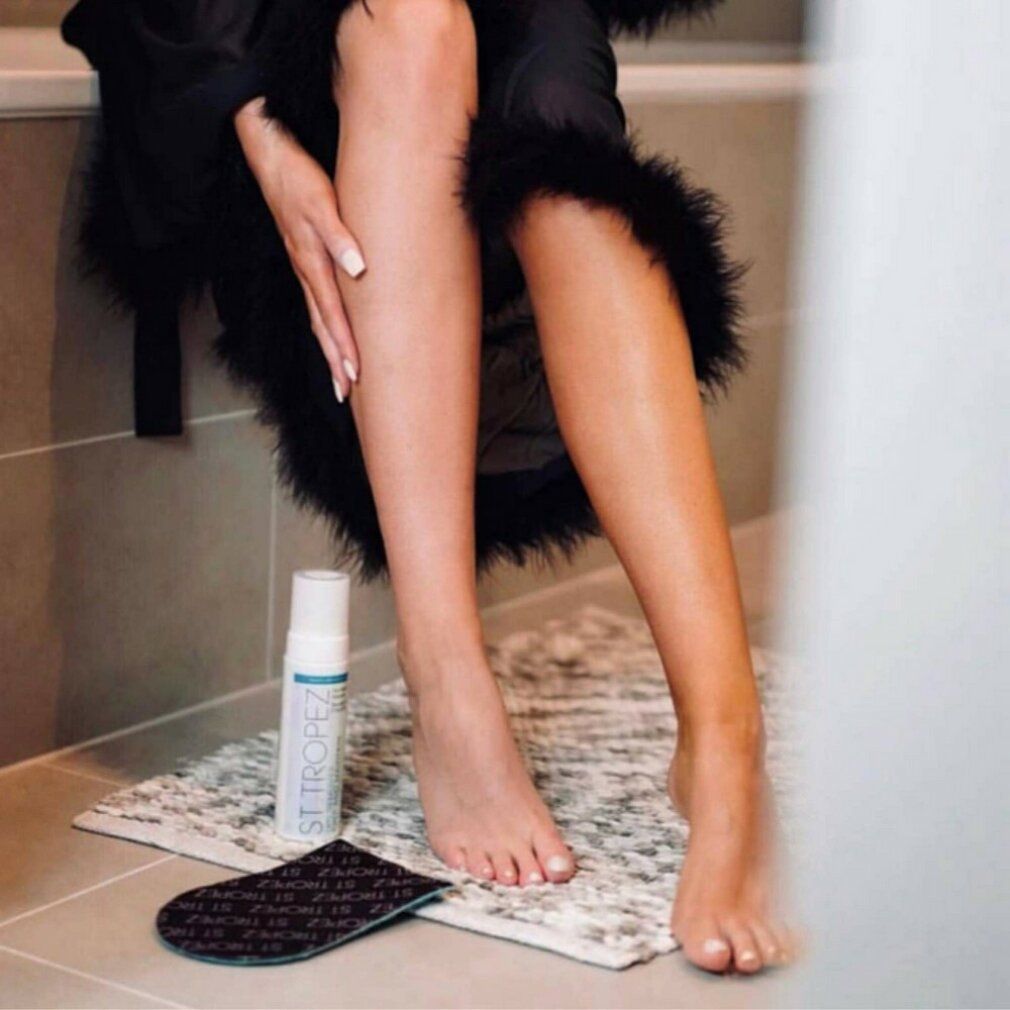 Persona seduta in bagno. Accanto a lei un prodotto e un applicatore. Gambe abbronzate e non.