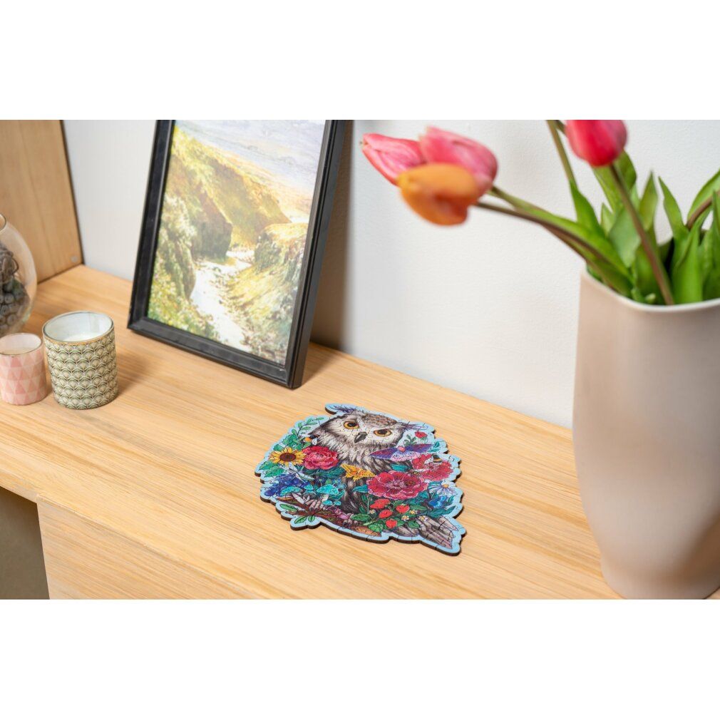 Puzzle finito con gufo e fiori su superficie in legno. Sullo sfondo: quadro, vaso con tulipani.