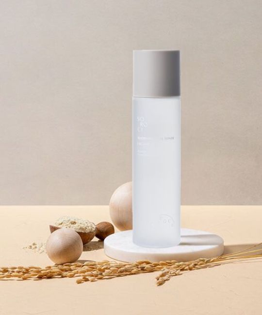 Flacone cilindrico con tappo grigio chiaro. Posizionato su una piattaforma bianca. Circondato da elementi decorativi.