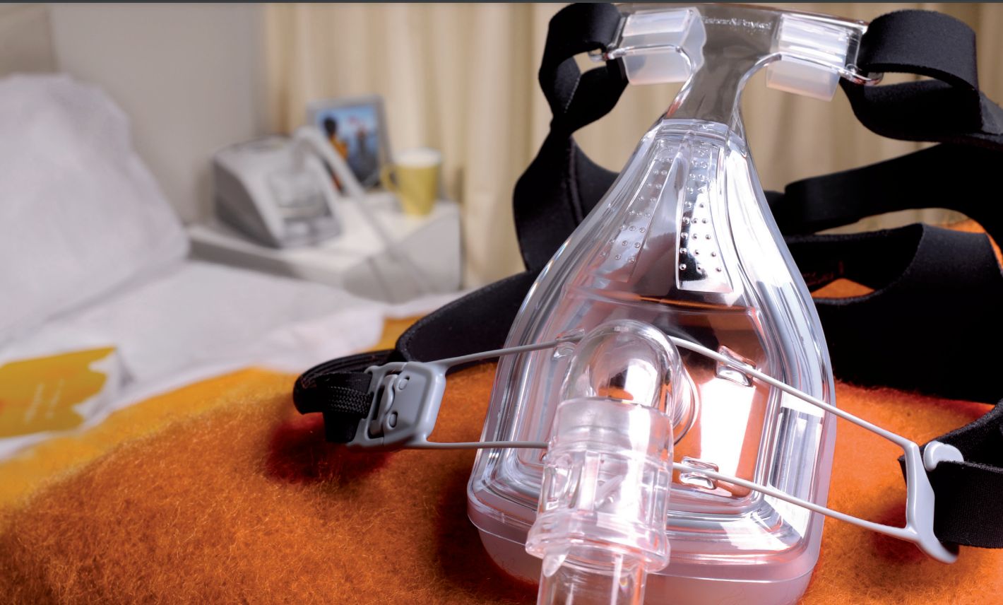 Maschera respiratoria con fascia su una coperta. Maschera trasparente, fascia nera. Sullo sfondo: letto, comodino.