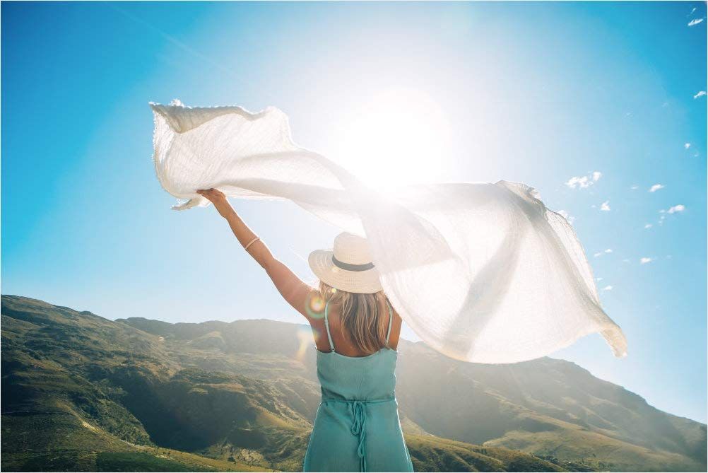 Donna con cappello e vestito, alza un tessuto bianco in aria. Sfondo: montagne e cielo blu con sole.