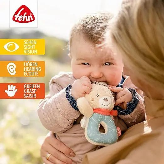Bambino tiene in bocca un sonaglio orsetto in peluche. Logo Fehn in alto a sinistra. Icone visive, uditive e tattili.