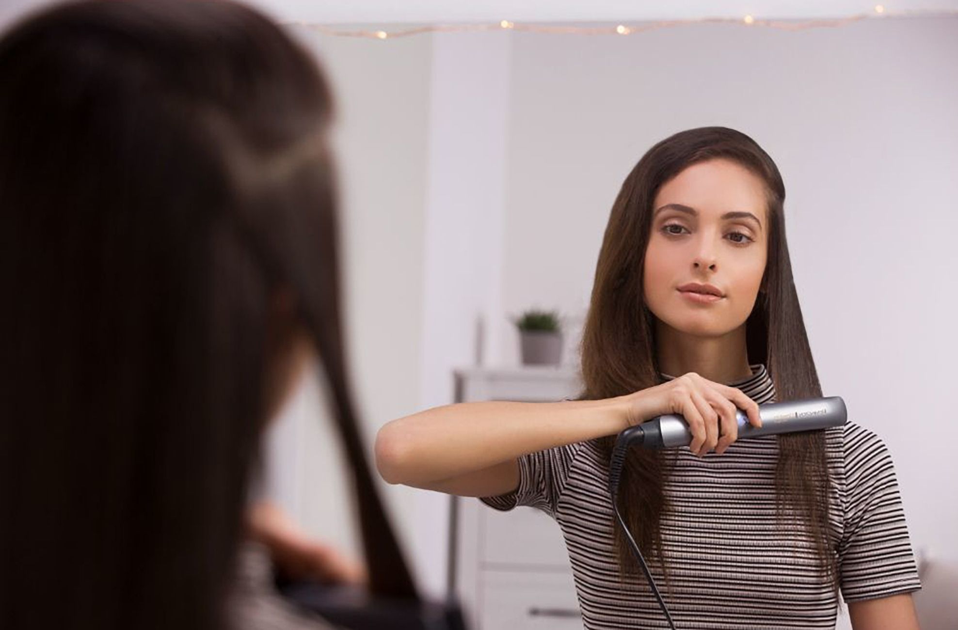Donna usa piastra per capelli davanti allo specchio. Capelli lisciati. Piastra grigia.