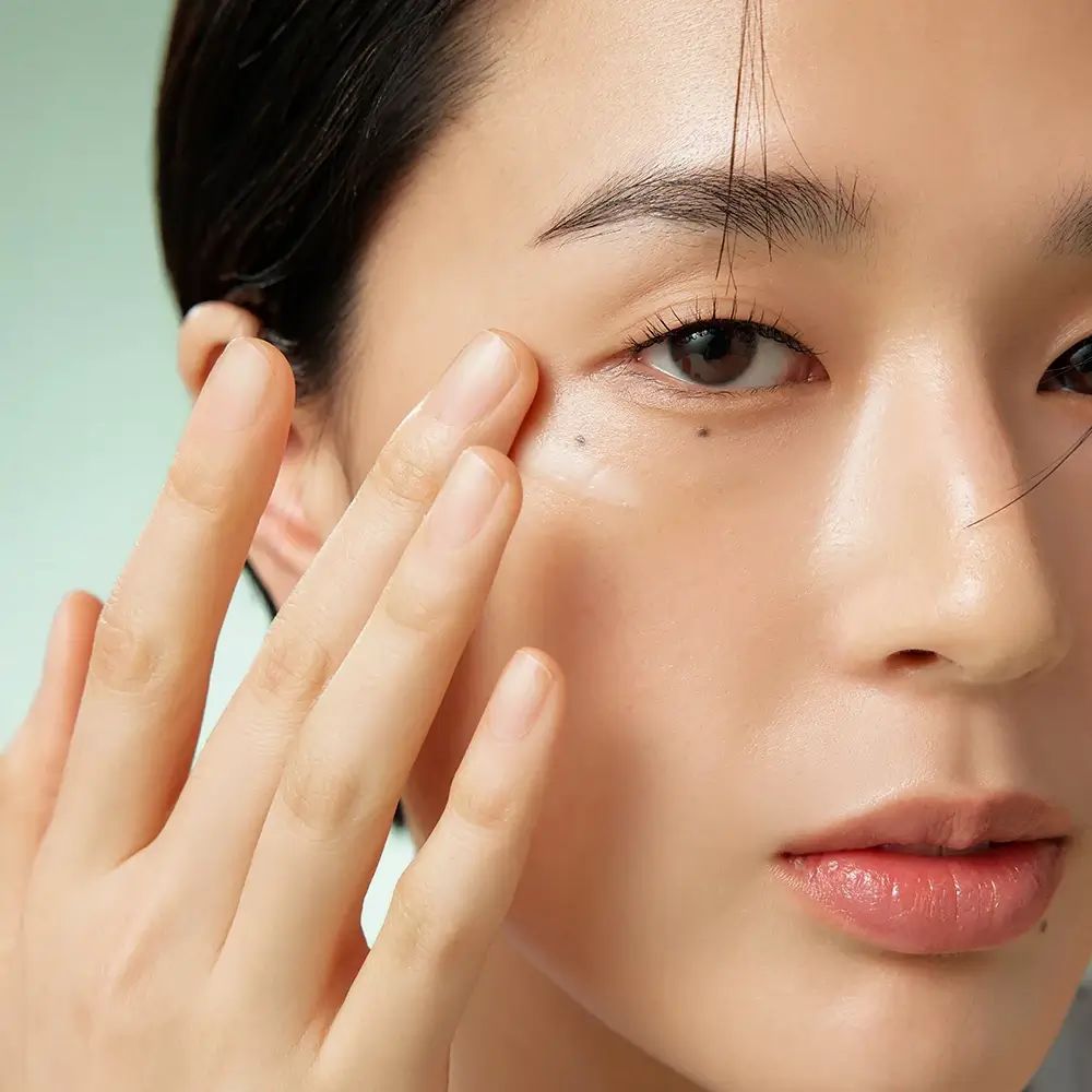 Primo piano di una persona che applica la crema sotto l'occhio. La mano tocca delicatamente la pelle. Pelle chiara, capelli scuri.