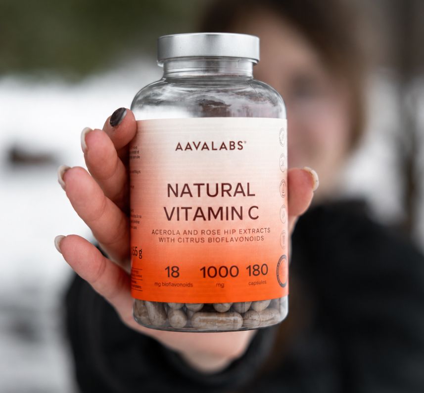 Mano che tiene un flacone di Aavalabs Vitamina C Naturale 1000 mg davanti a uno sfondo sfocato.