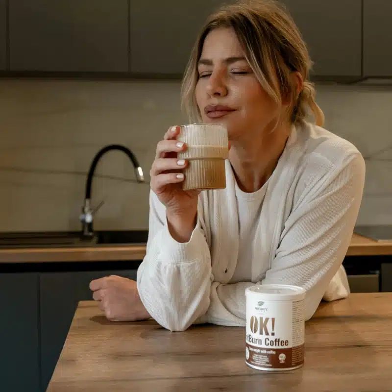 Una donna beve caffè accanto a una lattina OK! FatBurn Coffee al tavolo della cucina.