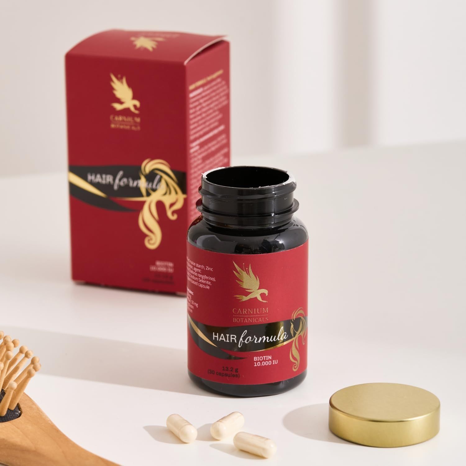 Flacone e scatola con etichetta rossa. Testo: CARNIUM BOTANICALS Hair Formula. Tappo dorato. Capsule accanto.