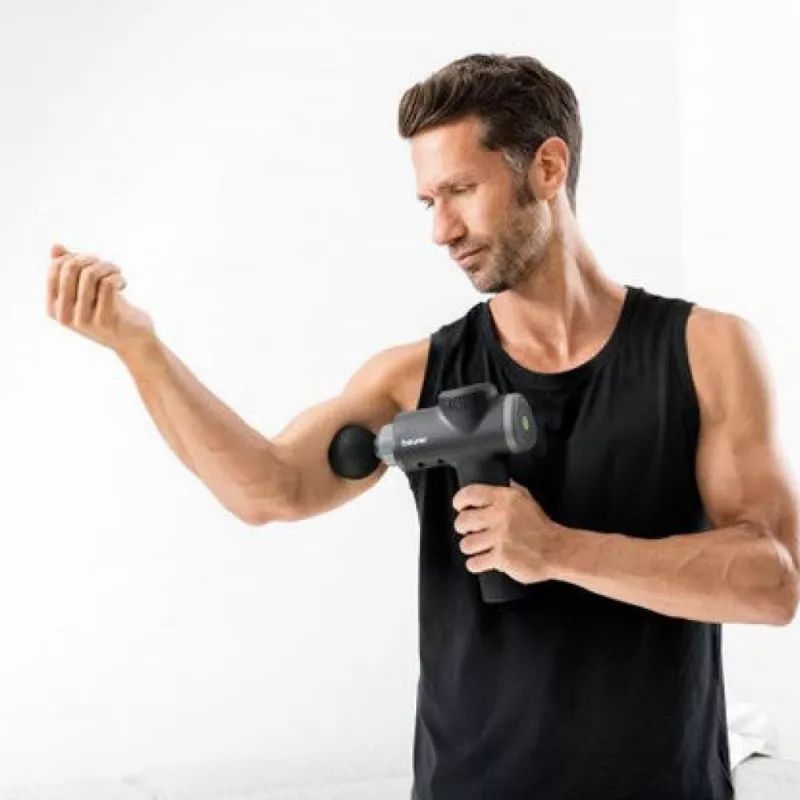 Uomo usa pistola massaggiante sul braccio. Pistola massaggiante nera con attacco grigio. Uomo indossa canotta nera. Sfondo bianco.