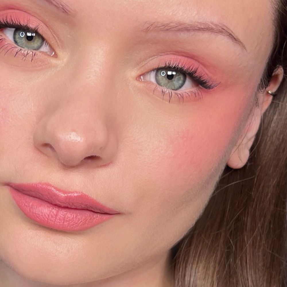 Primo piano di un viso con trucco rosa su occhi, guance e labbra. Occhi verdi.
