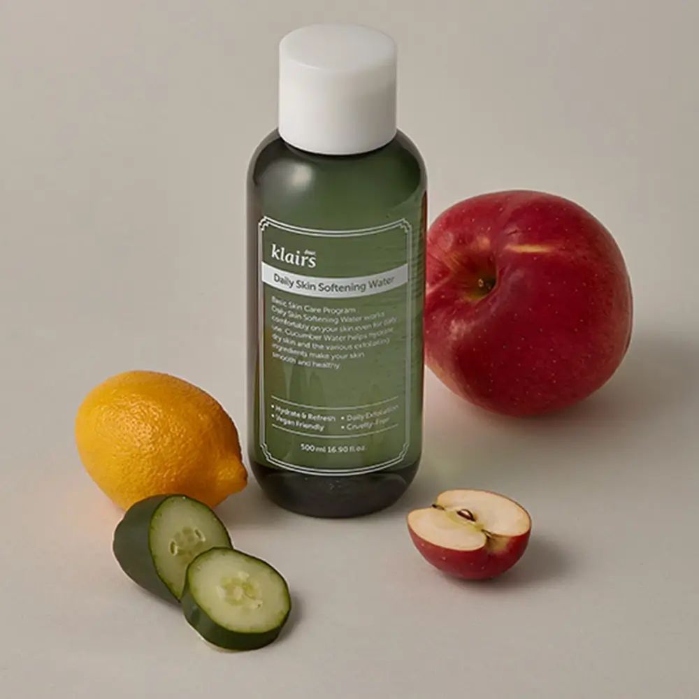 Flacone verde con etichetta prodotto, accanto limone, mela, fette di cetriolo. Su sfondo chiaro.