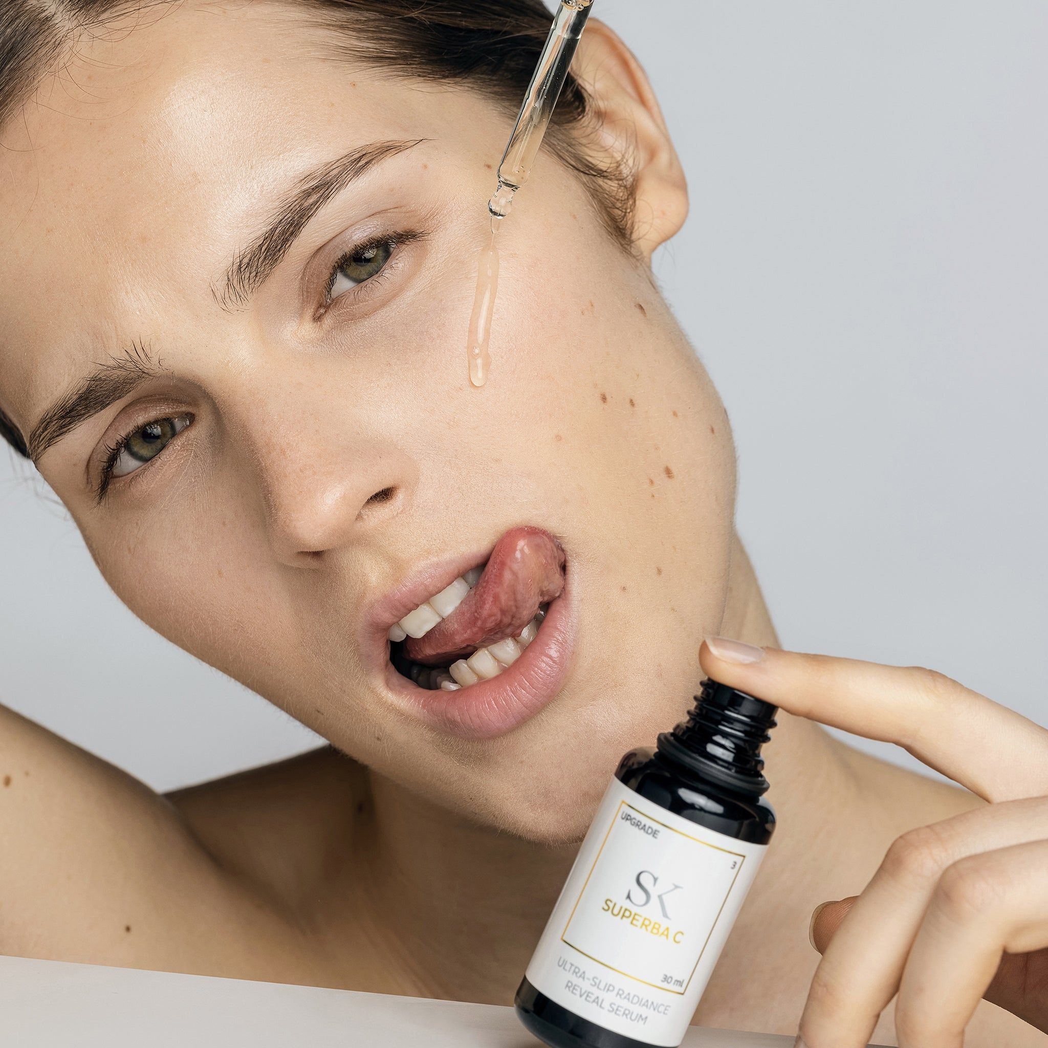 Donna con flacone e pipetta. Gocce sulla lingua. Il flacone indica "SK SUPERBA C" e "ULTRA-SLIP RADIANCE REVEAL SERUM".