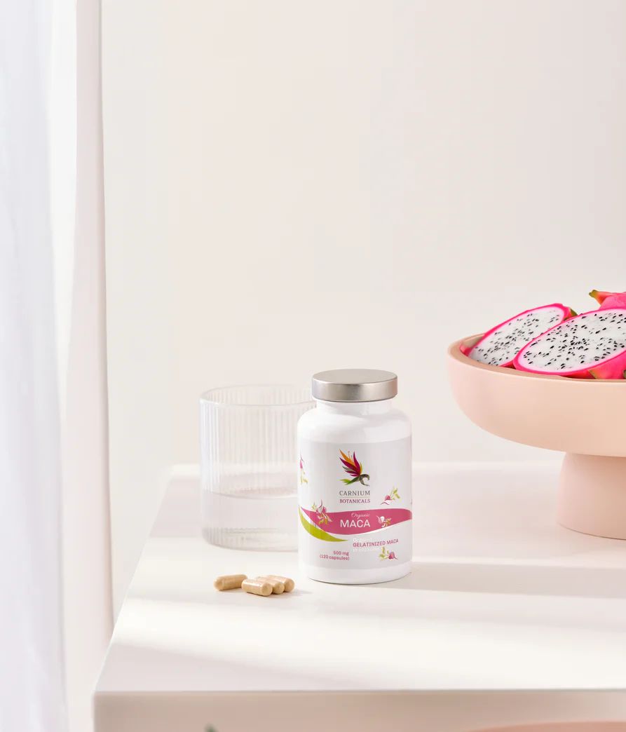 Flacone bianco con tappo argentato. Scritta: Carnium Botanicals, Maca, 500 mg (120 capsule). Bicchiere e coppa di frutta.