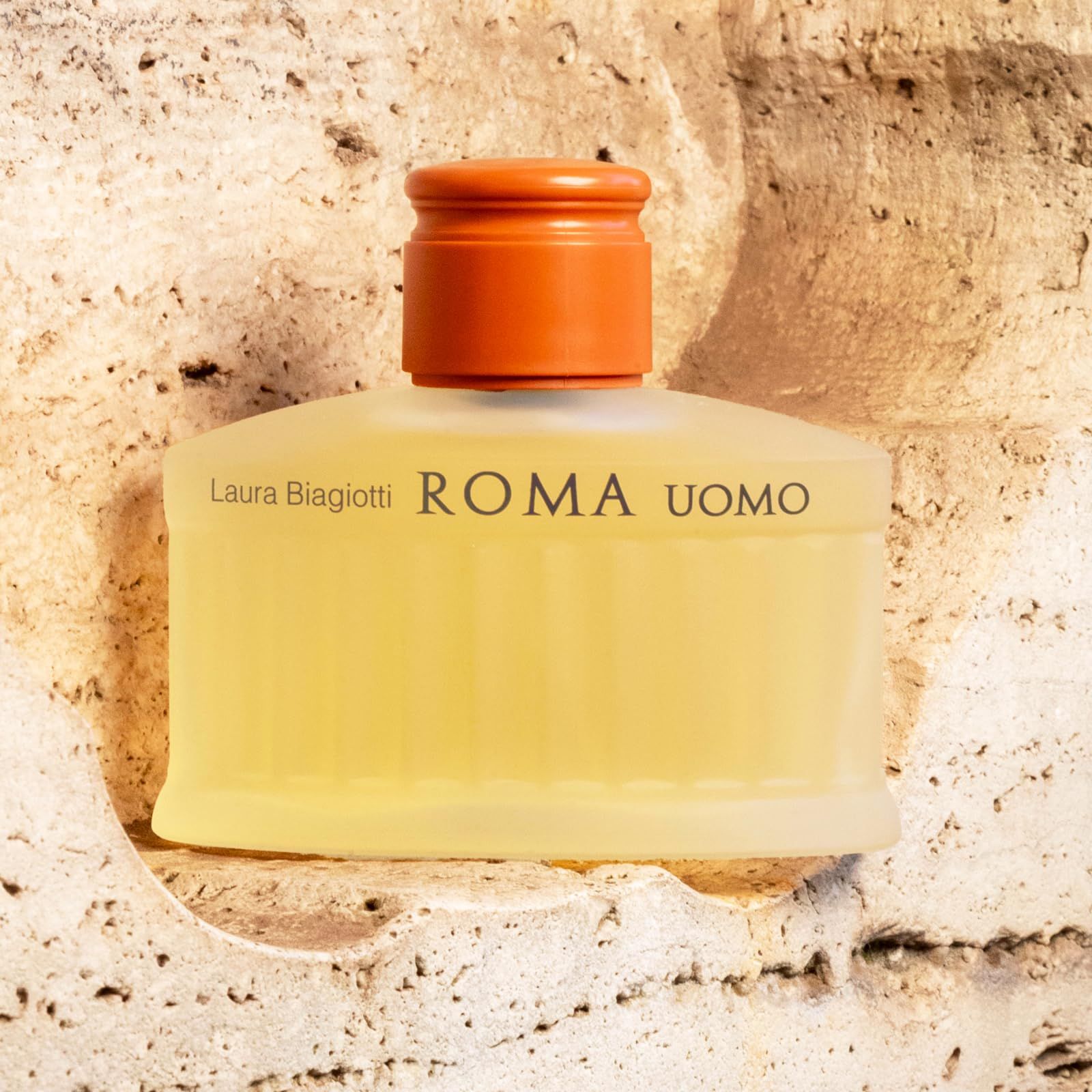 Flacone di profumo con liquido giallastro. Scritta: Laura Biagiotti ROMA UOMO. Su sfondo effetto pietra.
