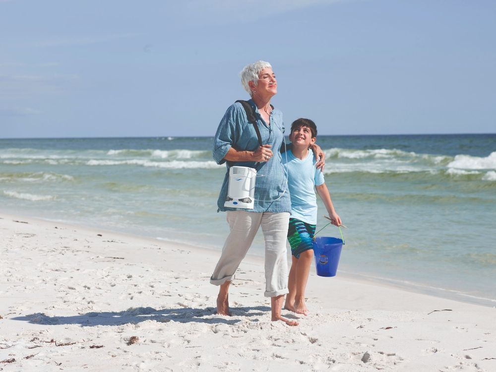 Donna anziana e bambino sulla spiaggia, entrambi con concentratore di ossigeno. La donna indossa il dispositivo. Spiaggia e mare sullo sfondo.