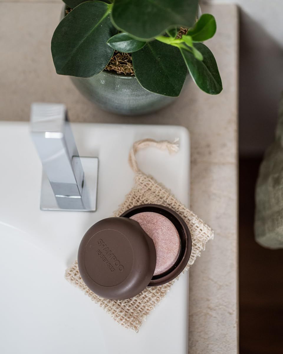 Contenitore rotondo marrone con shampoo solido rosa. Coperchio con scritta « Shampoo ». Su lavandino.