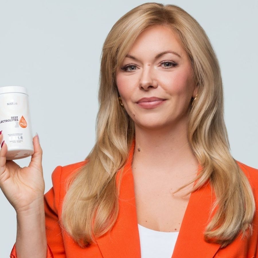 Donna tiene contenitore bianco BEST ELECTROLYTES. Indossa un blazer arancione. Capelli biondi.