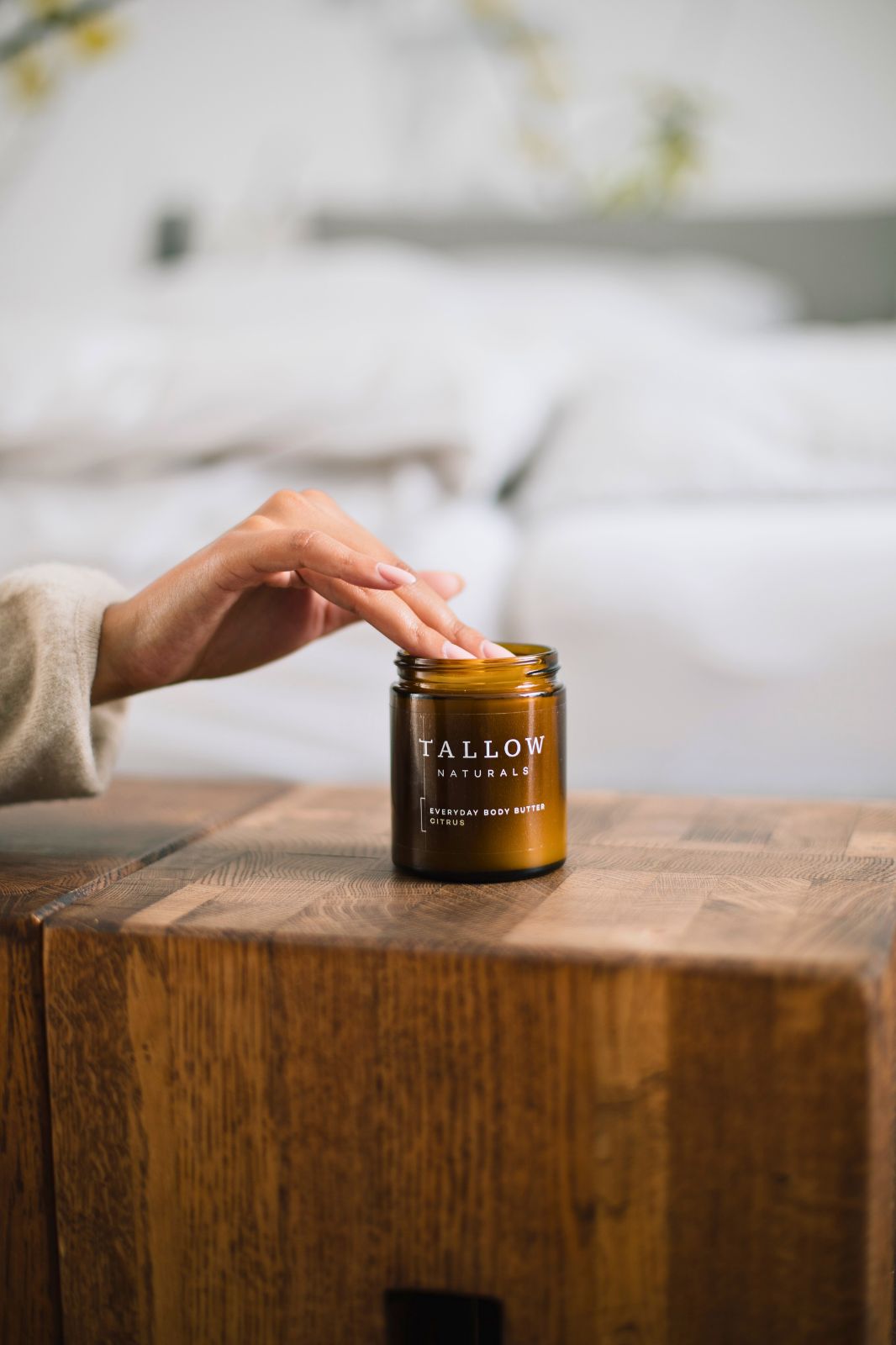Mano che tocca un barattolo marrone con coperchio nero. Scritta: TALLOW NATURALS, EVERYDAY BODY BUTTER CITRUS. Barattolo su legno.