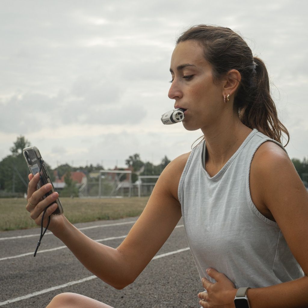 Donna con allenatore respiratorio in bocca, tiene smartphone. Su pista atletica.