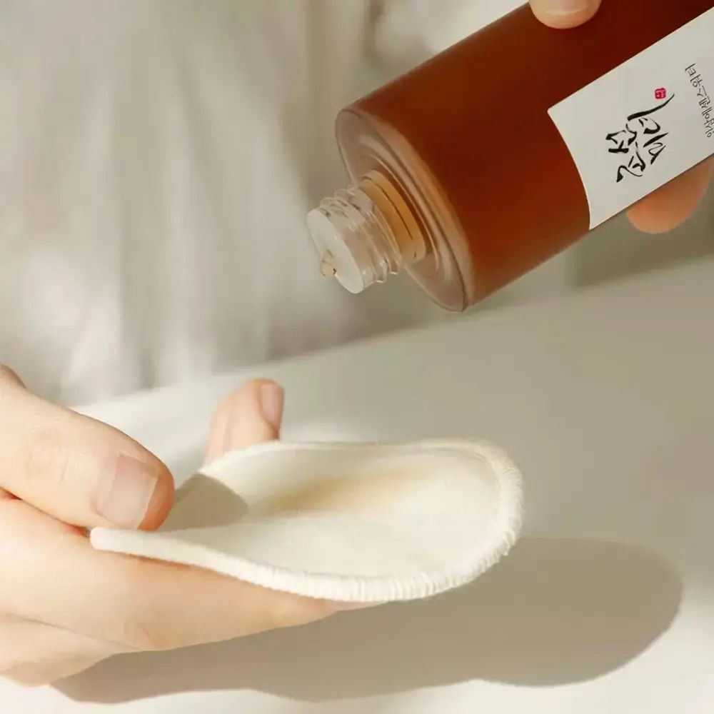 Mani che tengono un batuffolo di cotone imbevuto di liquido marrone. Bottiglia sullo sfondo. Scritta: Ginseng Essence Water.