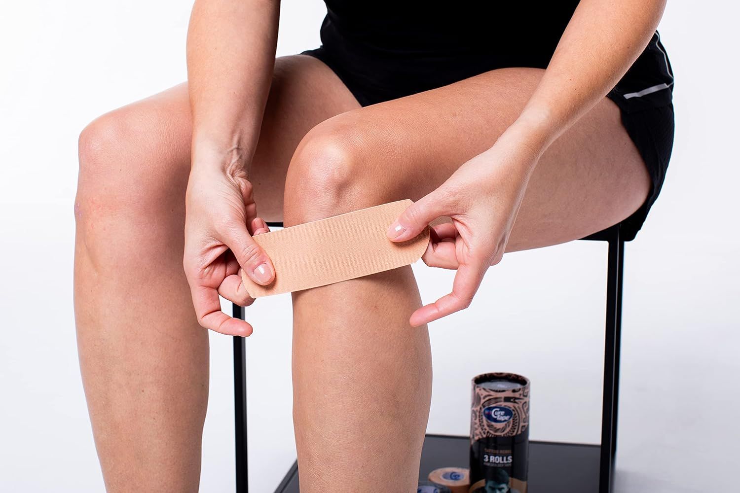 Persona applica nastro kinesiologico beige sul ginocchio. Rotolo di nastro a terra. Persona seduta su sgabello. Nastro applicato sulla gamba.