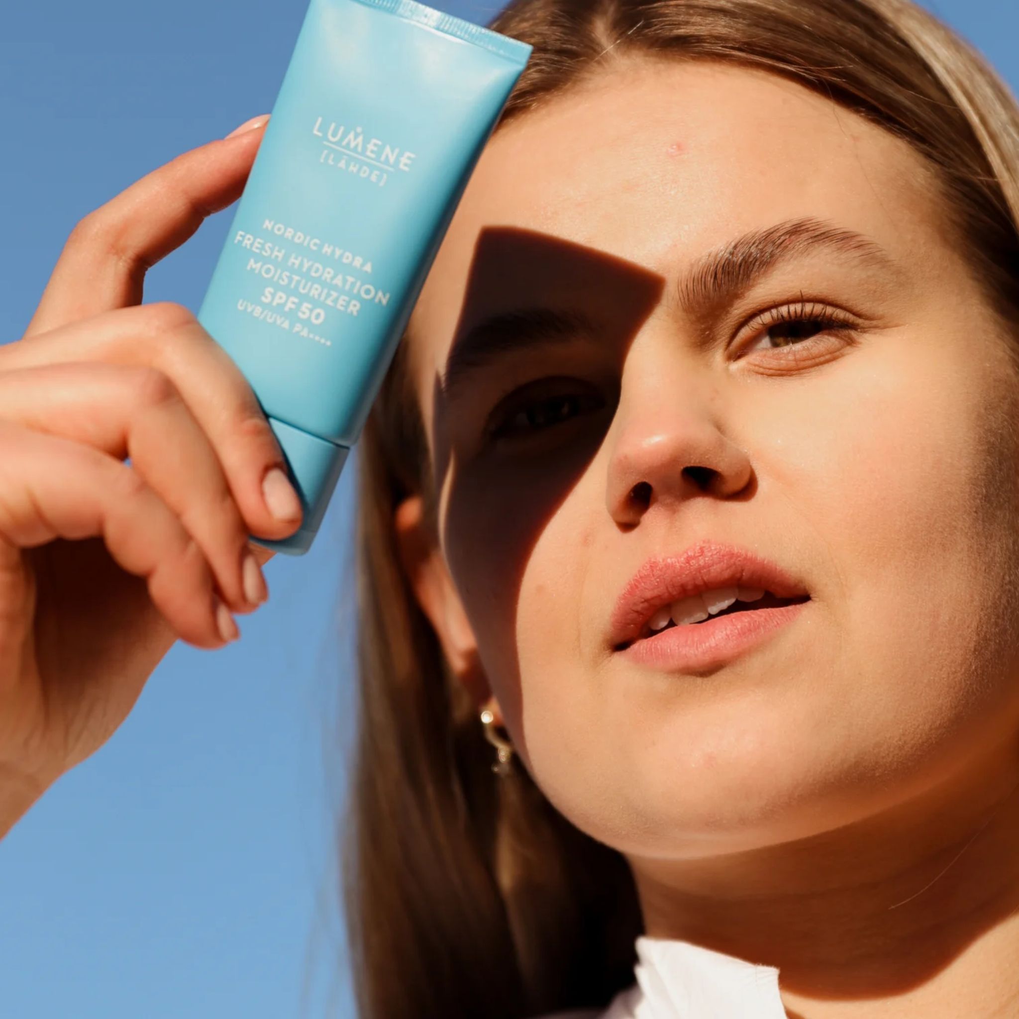 Donna tiene tubo azzurro chiaro davanti cielo blu. Scritto: LUMENE [LÄHDE], Nordic Hydra Fresh Hydration Moisturizer SPF50. Viso in ombra.