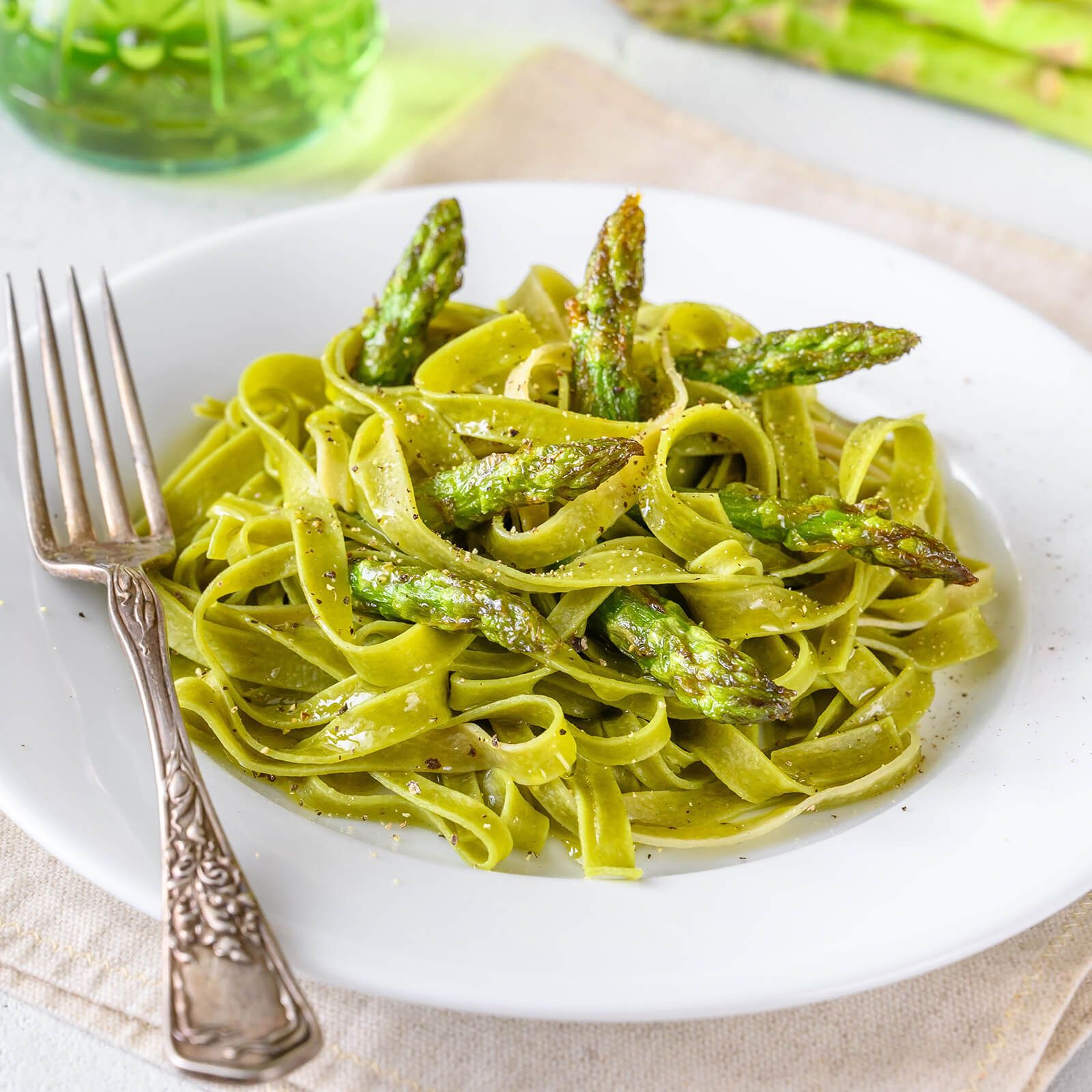 Piatto di tagliatelle verdi, asparagi e pepe. Su un panno beige, con una forchetta.