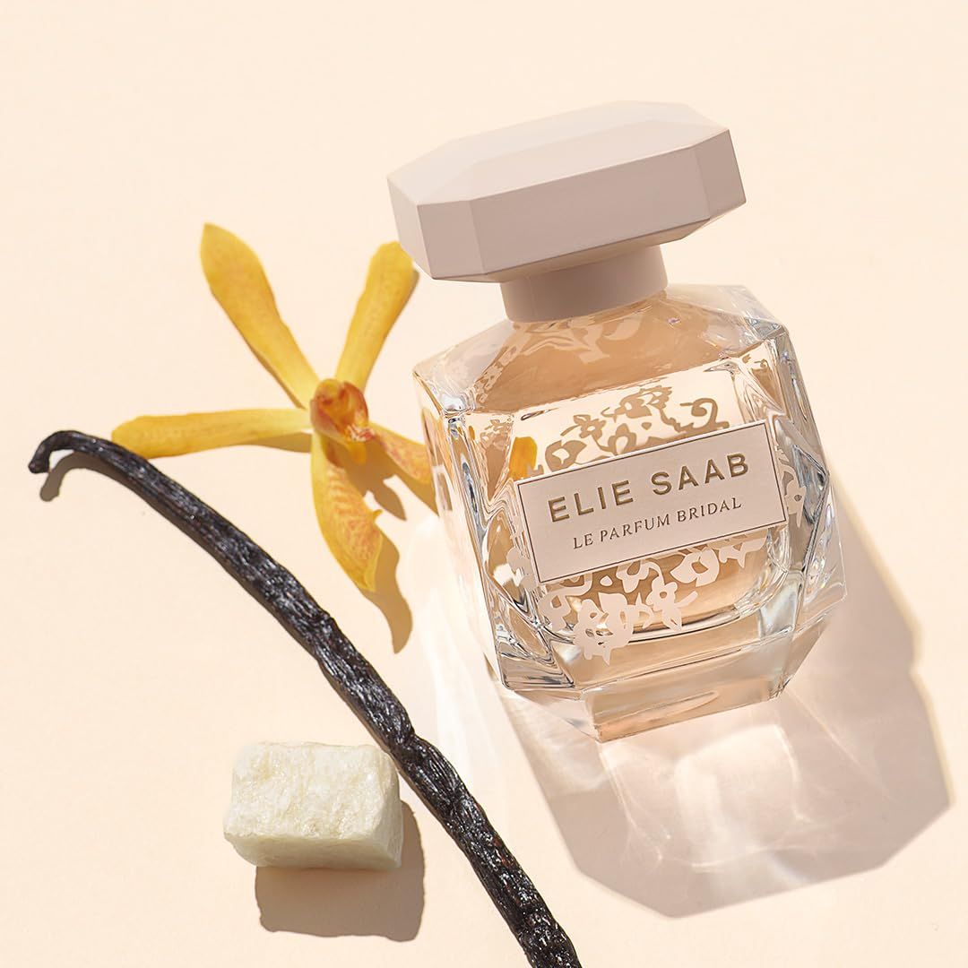 Flacone in vetro con tappo beige, ELIE SAAB Le Parfum Bridal. Baccello di vaniglia, fiore e cubo accanto.