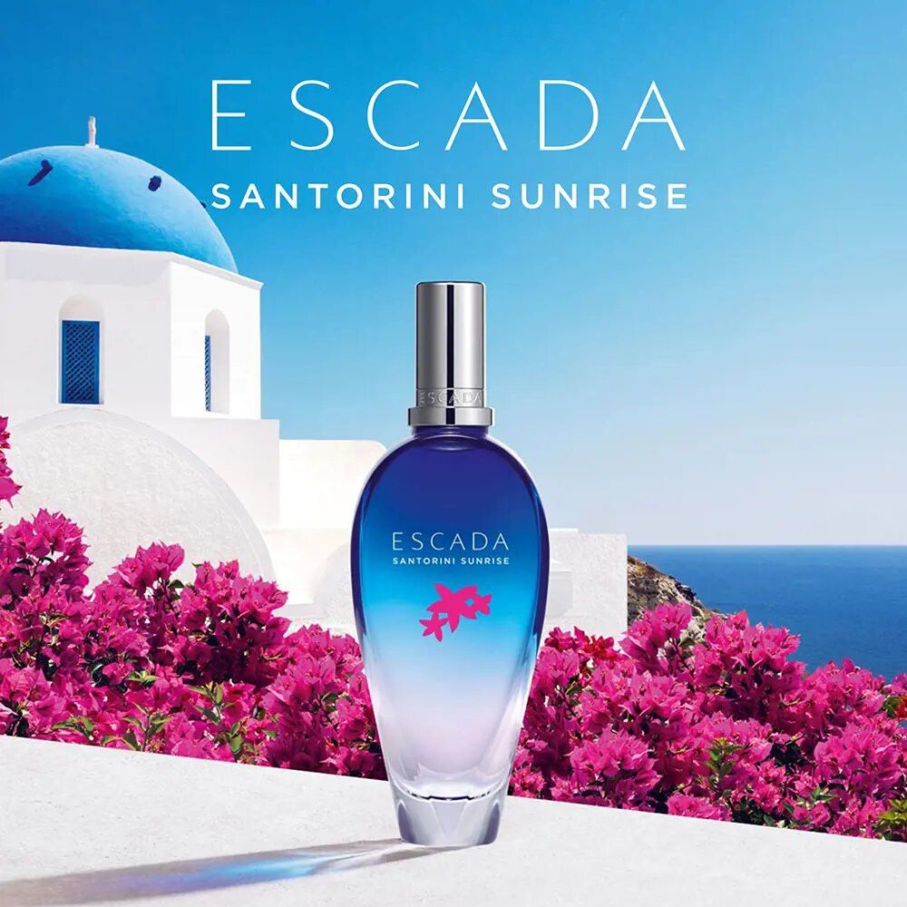 Flacone blu trasparente davanti a uno sfondo greco. Scritta: ESCADA SANTORINI SUNRISE. Fiori rosa.
