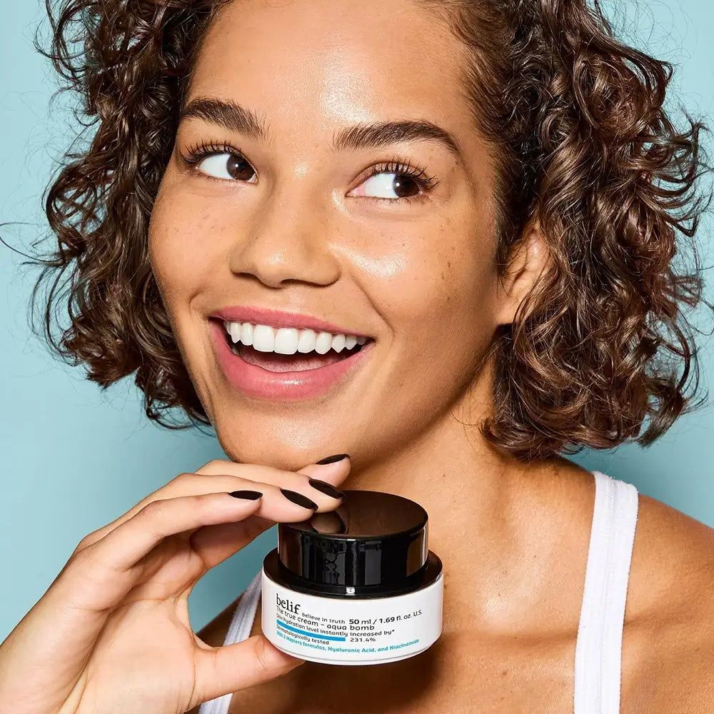 Donna sorridente con capelli ricci che tiene un vasetto di Belif The True Cream Aqua Bomb.