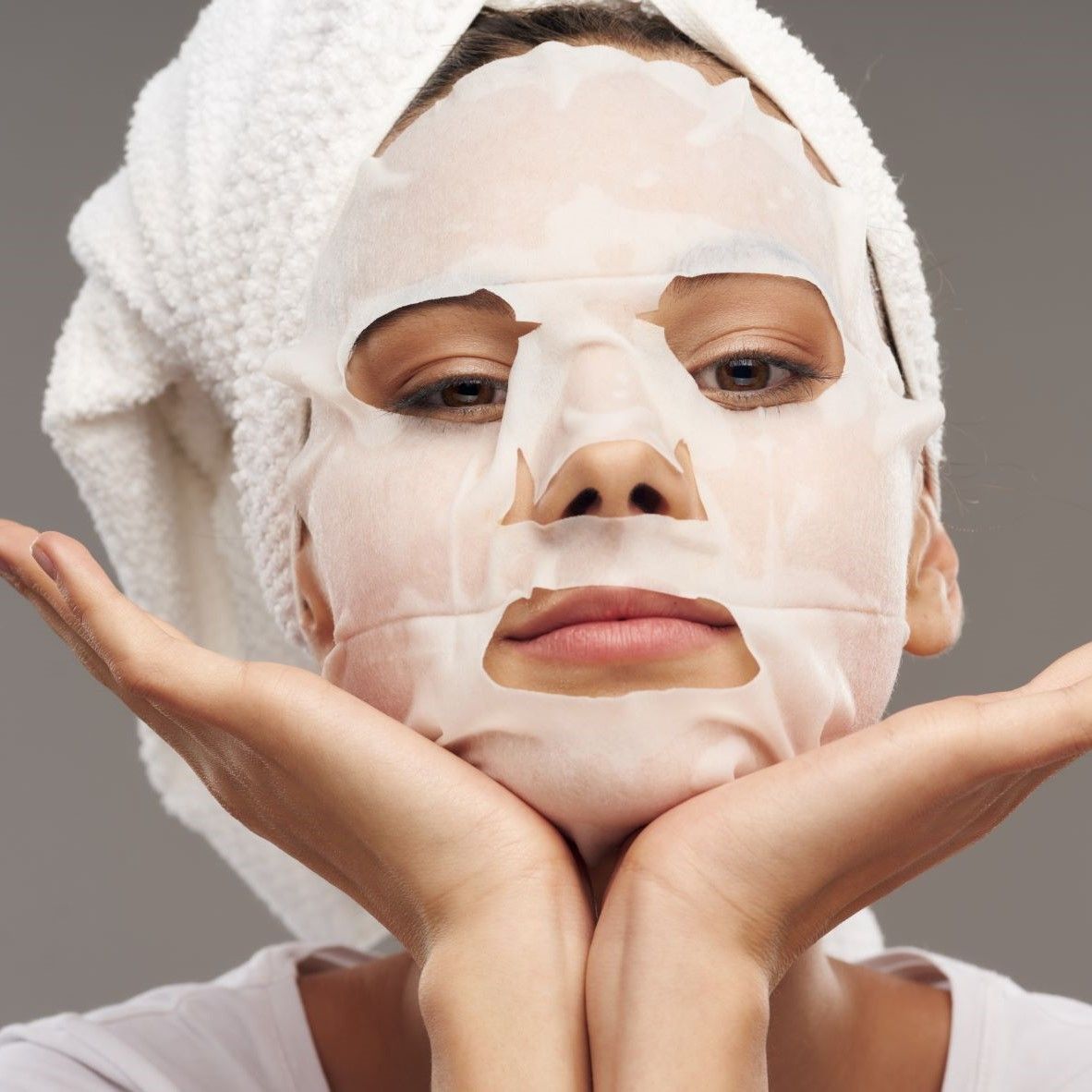 Maschera per il viso sul viso di una donna. Donna con asciugamano. La maschera copre il viso. Occhi, naso, bocca visibili. Mani sotto il mento.