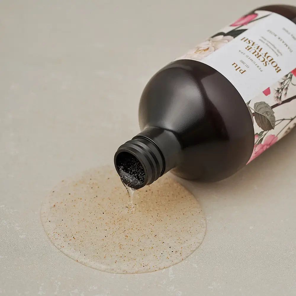 PLU Scrub Body Wash Damask Rose versato da un flacone marrone su una superficie.