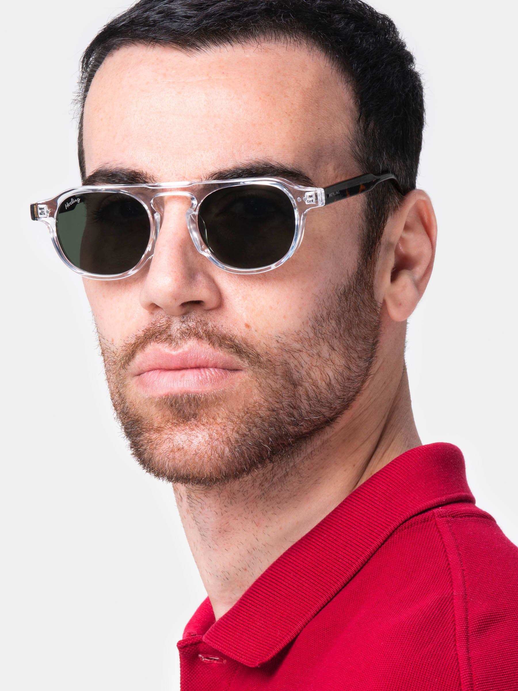 Uomo con occhiali da sole con montatura trasparente e lenti scure. Logo Herling sulla lente. Polo rosso.