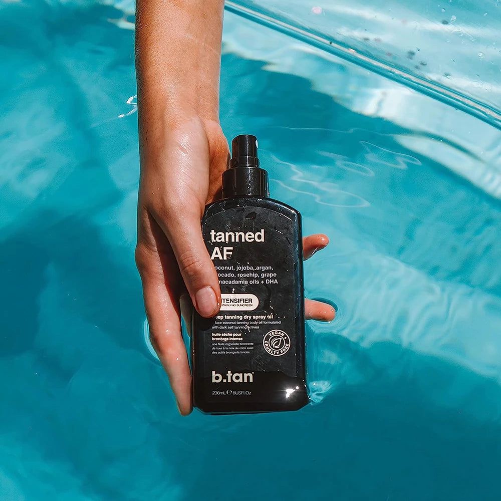 Flacone spray nero 'tanned AF' in una mano sopra l'acqua blu. 'b.tan' è visibile. Contiene cocco, jojoba, argan, avocado, rosa canina, uva e oli di macadamia + DHA.