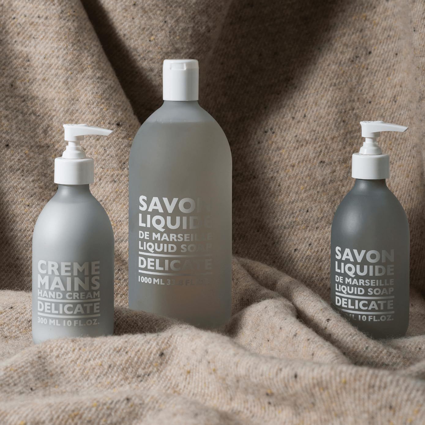 Tre flaconi su tessuto beige. Due con pompa, uno senza. Testo: Savon Liquide Delicate, Crème Mains Delicate.