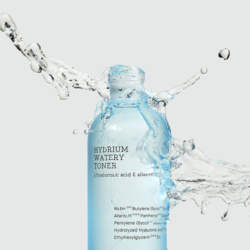Flacone di toner per la pelle, liquido blu, tappo bianco. L'acqua schizza sul flacone. Scritte: HYDRIUM WATERY TONER, ingredienti.