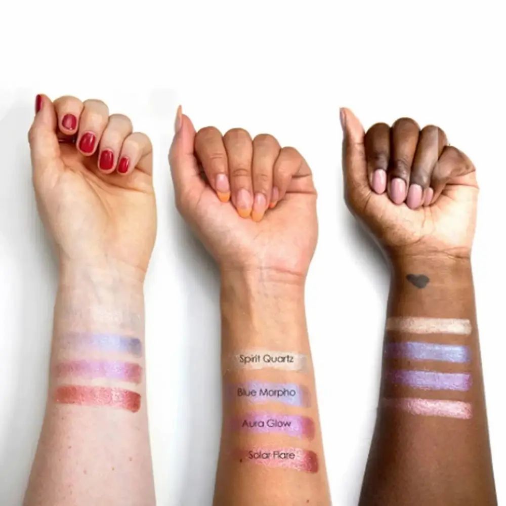 Tre braccia con diverse tonalità di pelle mostrano campioni di ombretto. I nomi dei colori sono etichettati: Spirit Quartz, Blue Morpho, Aura Glow, Solar Flare.