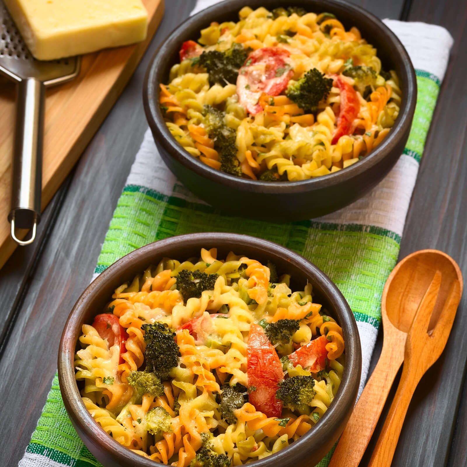 Due ciotole di fusilli tricolore al forno con broccoli e pomodori. Su fondo in legno.
