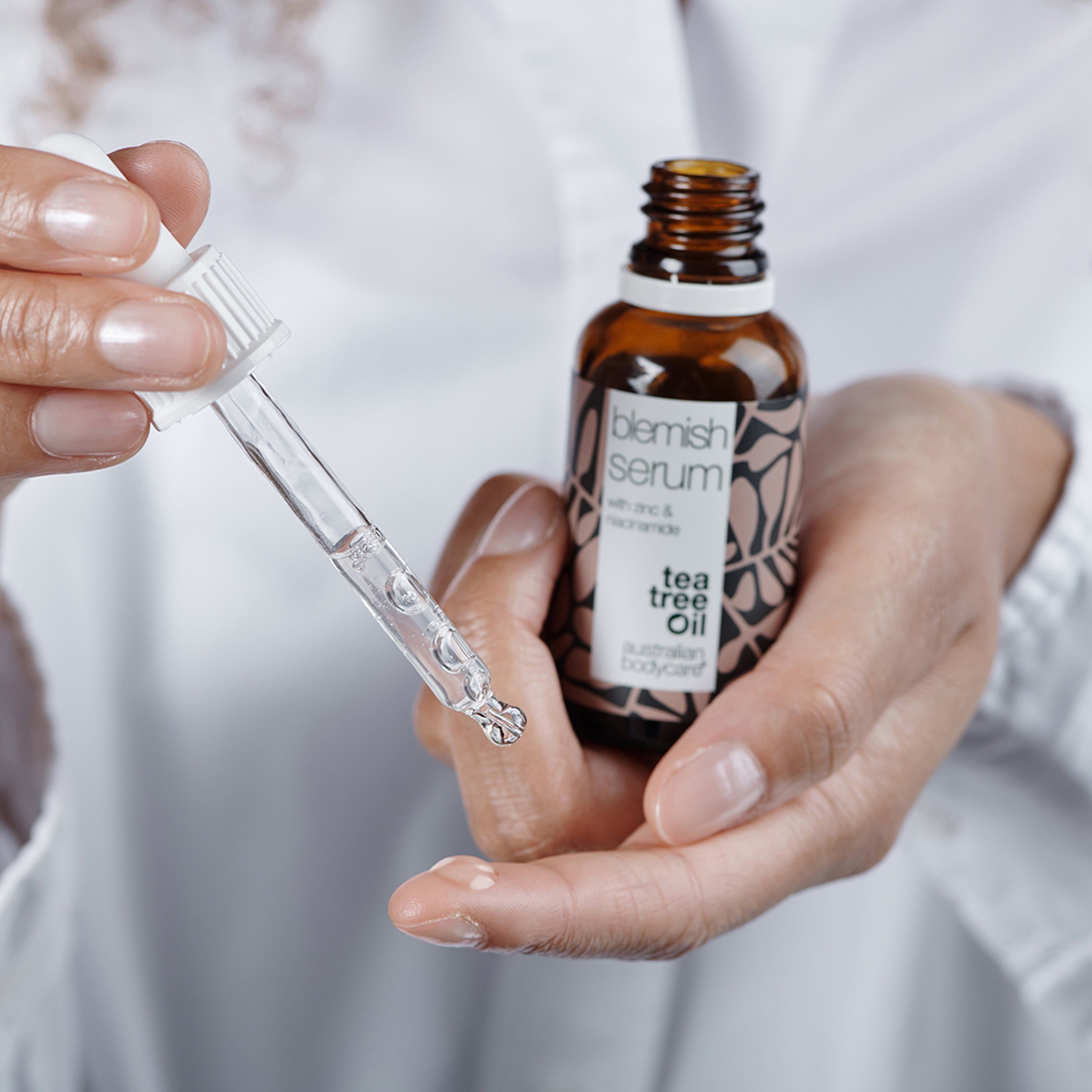 Mani che tengono flacone di siero e pipetta. Scritta: Blemish Serum, Tea Tree Oil, Australian Bodycare.