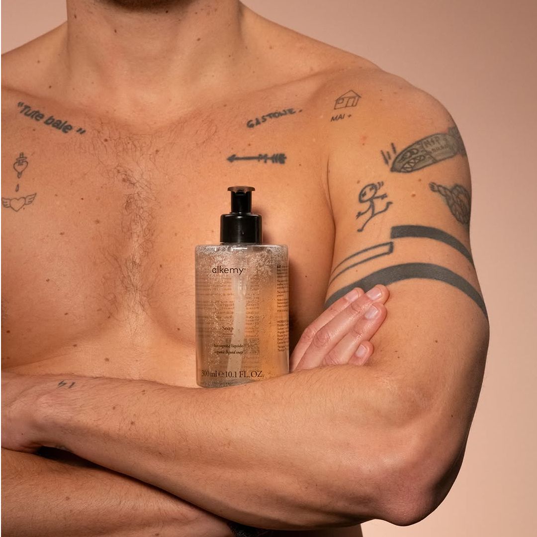 Uomo tatuato tiene flacone di sapone liquido trasparente sul petto.