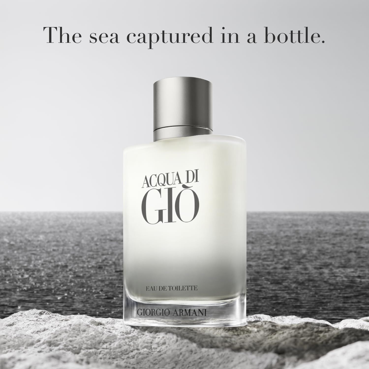 Flacone in vetro con tappo argentato. Scritta "ACQUA DI GIÒ" e "GIORGIO ARMANI". Flacone su sabbia di fronte al mare.