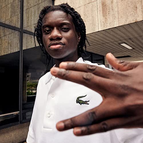 Un giovane con trecce indossa una polo bianca con logo coccodrillo verde. Un edificio sullo sfondo.