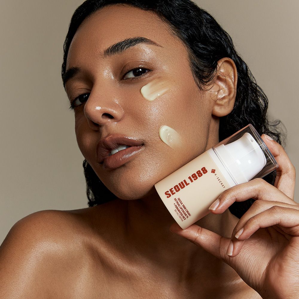 Una donna con la crema viso K-Secret Seoul 1988 sulla pelle e un flacone dispenser in mano.