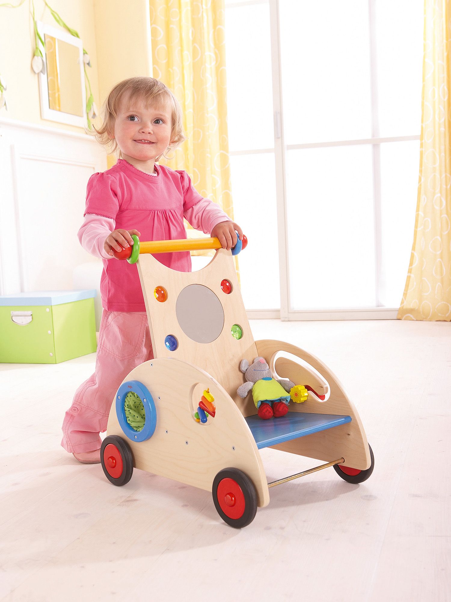 Bambino piccolo dietro un girello in legno. Il girello ha ruote, un manico e dei giocattoli.