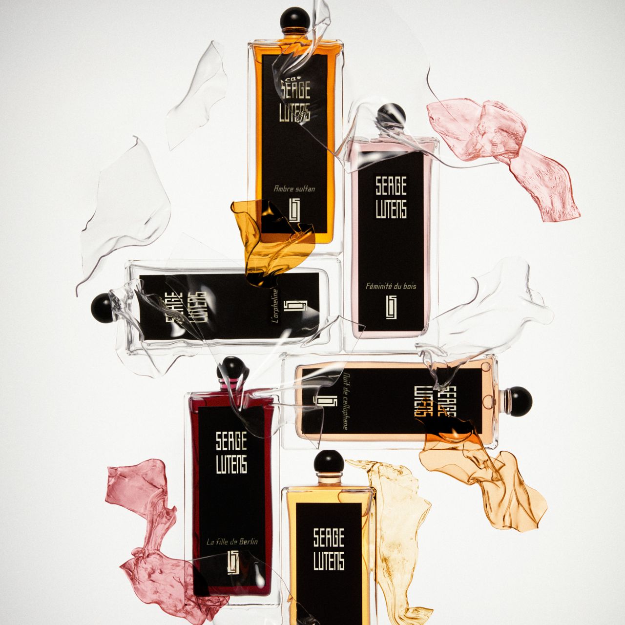 Diversi flaconi Serge Lutens, circondati da vetro rotto e tessuti colorati. Diverse fragranze.