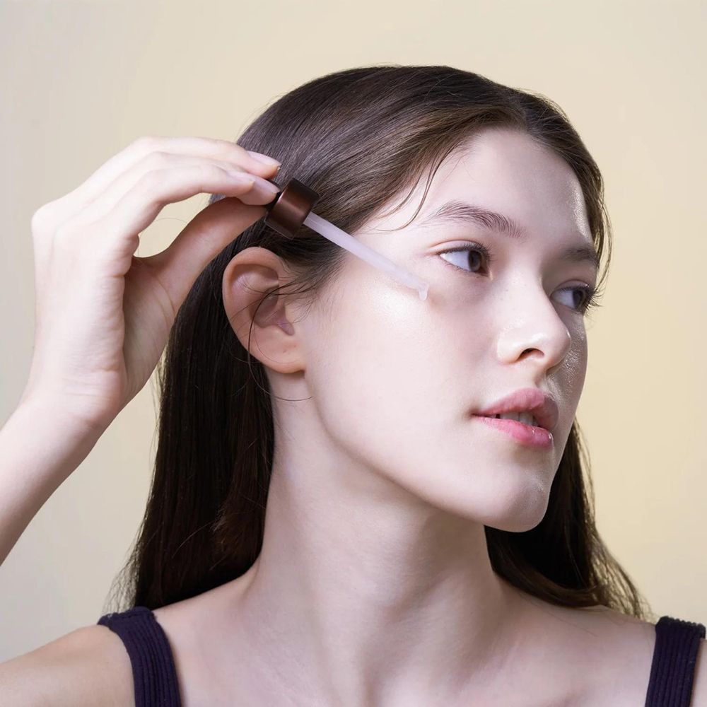 Giovane donna con capelli lunghi che applica siero sul viso con una pipetta. Pelle chiara, sguardo laterale.
