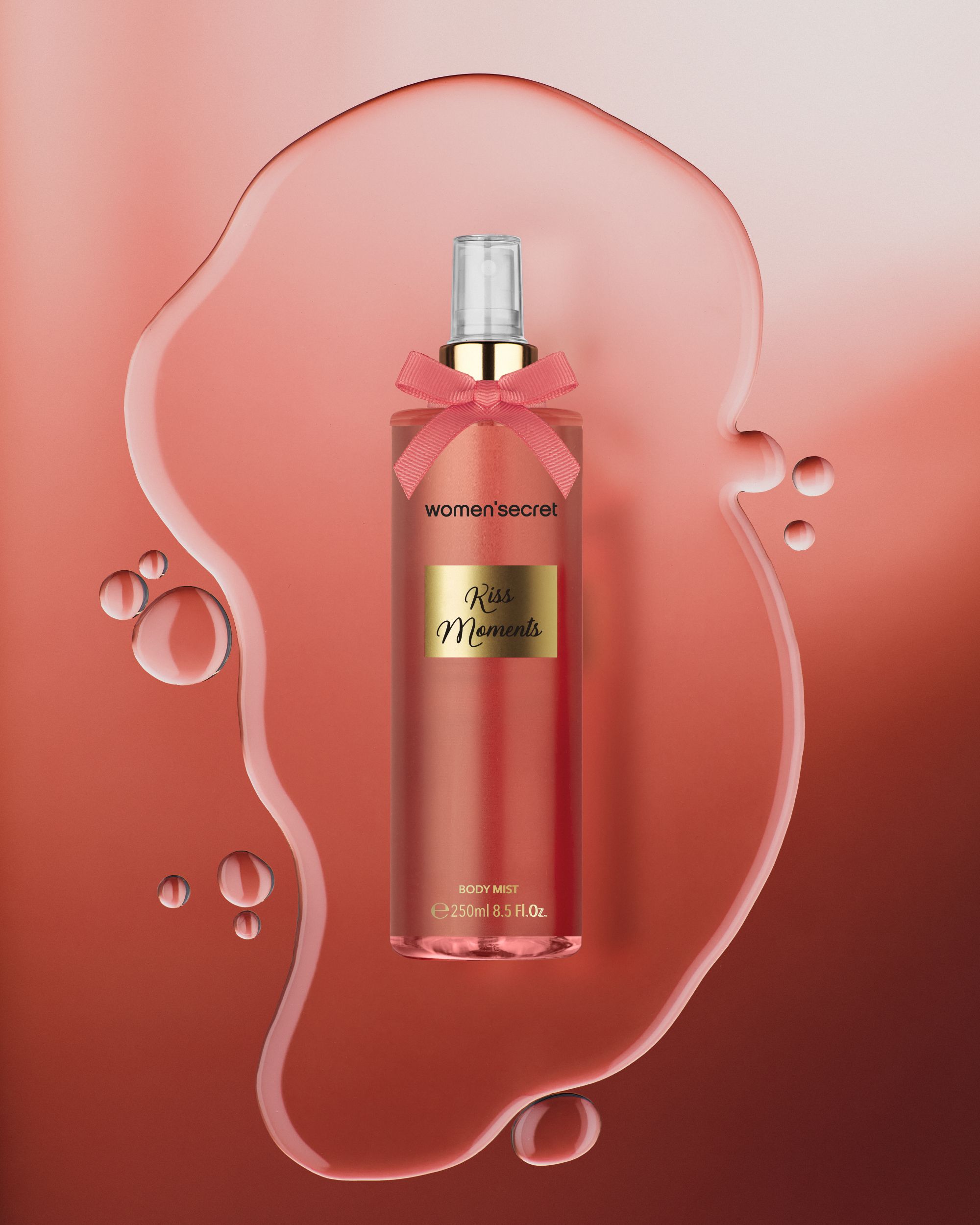 Flacone di Body Mist con fiocco rosa. Scritta: Women'secret, Kiss Moments. Etichetta dorata. Su sfondo rosa con gocce d'acqua.