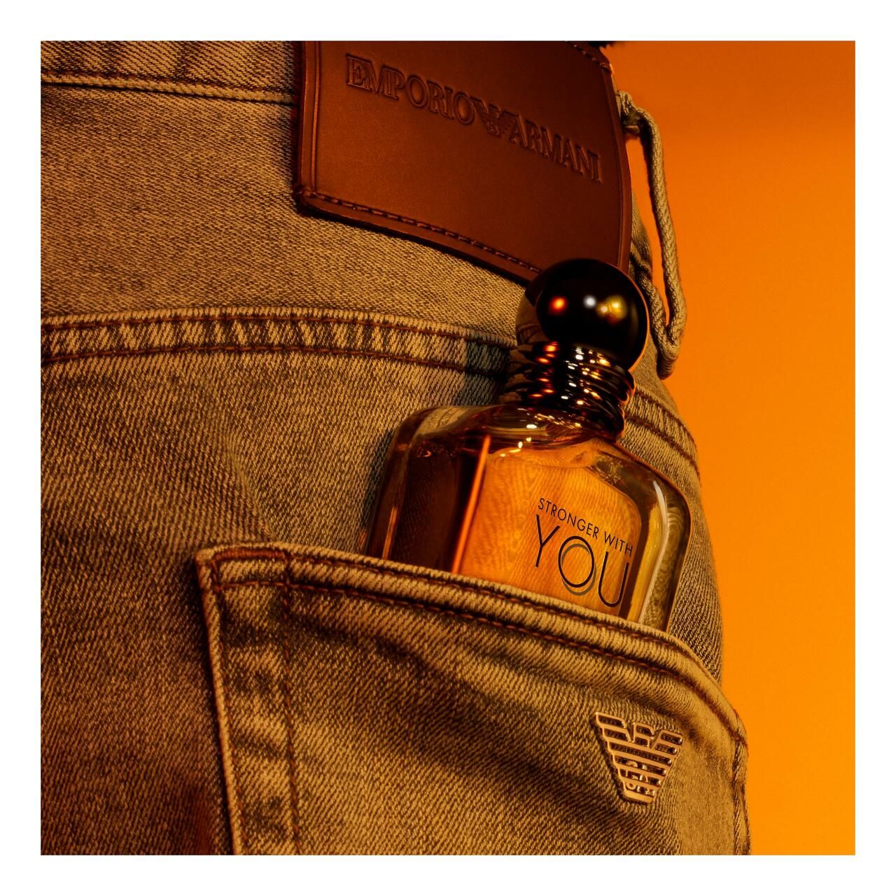 Flacone in vetro in tasca di jeans. Scritta: STRONGER WITH YOU e EMPORIO ARMANI. Etichetta in pelle con logo.