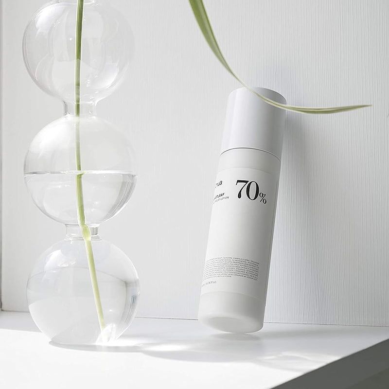 Flacone di lozione bianco vicino a vasi di vetro e pianta. Scritta: Anua, Heartleaf 70% Daily Releaf Lotion, 70%.