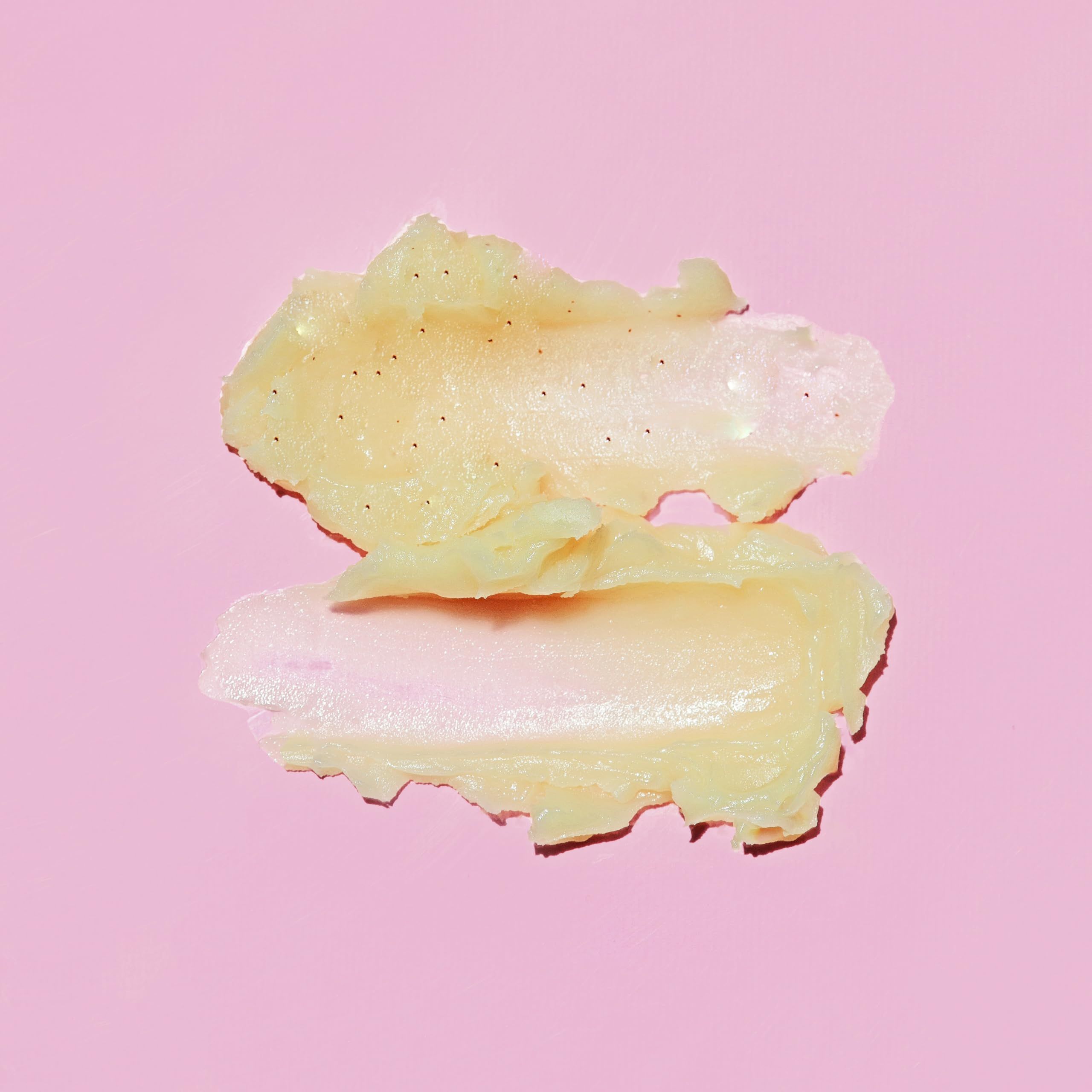 Due strisce di scrub labbra color crema su sfondo rosa. Particelle nere visibili.